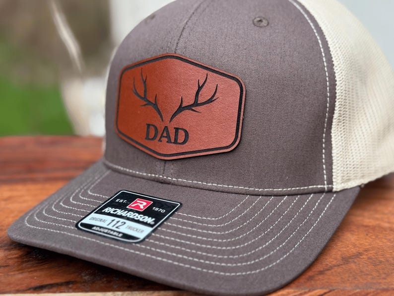 Pu&ograve; includere: Un cappellino trucker grigio antracite e beige con una toppa in pelle marrone sul davanti. La toppa mostra una grafica nera di corna di cervo sopra la scritta DAD. Un adesivo nero sulla visiera riporta la scritta Richardson Original 112 Trucker.
