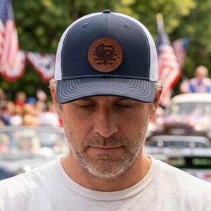 May include: A navy blue and white trucker hat featuring a circular brown leather patch on the front. The patch displays an eagle emblem, the text United States of America, and the text 250th Anniversary with a striped flag design in the center.