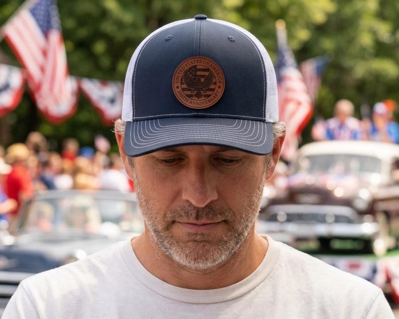 May include: A navy blue and white trucker hat featuring a circular brown leather patch on the front. The patch displays an eagle emblem, the text United States of America, and the text 250th Anniversary with a striped flag design in the center.