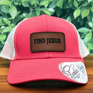 May include: A red trucker hat with a white mesh back and a curved brim. The hat features a brown leather patch with the words "FIND JESUS" in bold, black letters. A white tag with the text "Evolve Elude" is attached to the brim.