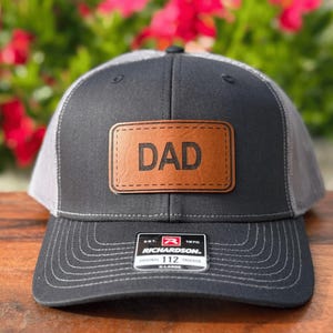 May include: A black and gray trucker hat with a brown leather patch that reads "DAD". The hat has a curved brim and a mesh back. The Richardson 112 hat is X-Large.