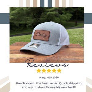 May include: A light gray baseball cap with a white mesh back and a brown rectangular leather patch on the front. The patch features the word Daddy in a black cursive script. A small black sticker is visible on the brim of the cap.
