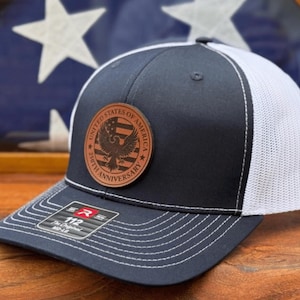 May include: A navy blue and white trucker hat featuring a circular brown leather patch on the front. The patch displays an eagle graphic, the text United States of America, and 250th Anniversary. A sticker on the brim reads 112 The Original MD-LG.