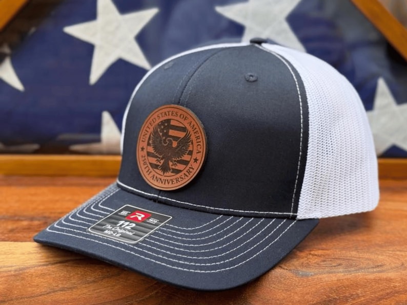 May include: A navy blue and white trucker hat featuring a circular brown leather patch on the front. The patch displays an eagle graphic, the text United States of America, and 250th Anniversary. A sticker on the brim reads 112 The Original MD-LG.