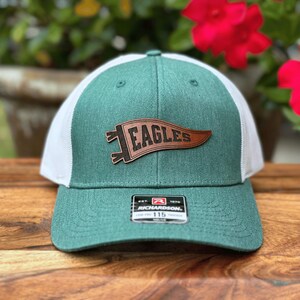 May include: A green and white trucker hat with a brown leather patch that reads "EAGLES". The hat has a curved brim and a mesh back. The Richardson 115 label is visible on the front.