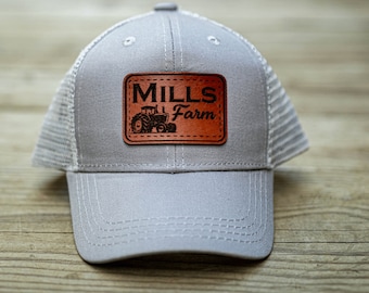 Peut inclure: Un chapeau de camionneur gris et blanc avec un patch en cuir marron qui dit "MILLS Farm" avec un graphique de tracteur.