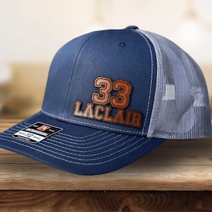 May include: A navy blue and gray trucker hat with a brown leather patch that says "33 LACLAIR" in white lettering.