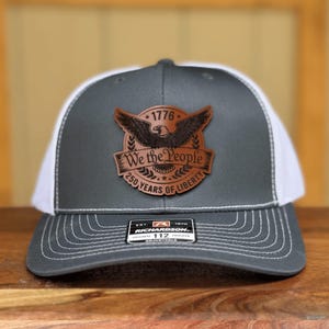 May include: A charcoal grey and white trucker hat featuring a brown leather patch on the front. The patch displays an eagle, the text 1776, We the People, and 250 YEARS OF LIBERTY. A black sticker on the brim reads RICHARDSON 112 TRUCKER.