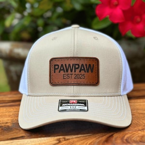 May include: A beige and white trucker hat with a brown leather patch that says "PAWPAW EST 2025".