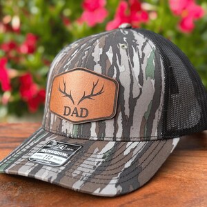 Pu&ograve; includere: Un cappellino trucker con motivo mimetico nei toni del marrone, grigio e verde e una parte posteriore in rete nera. Una toppa color cuoio sul davanti mostra un'illustrazione di corna di cervo sopra la scritta DAD. Un adesivo &egrave; presente sulla visiera.