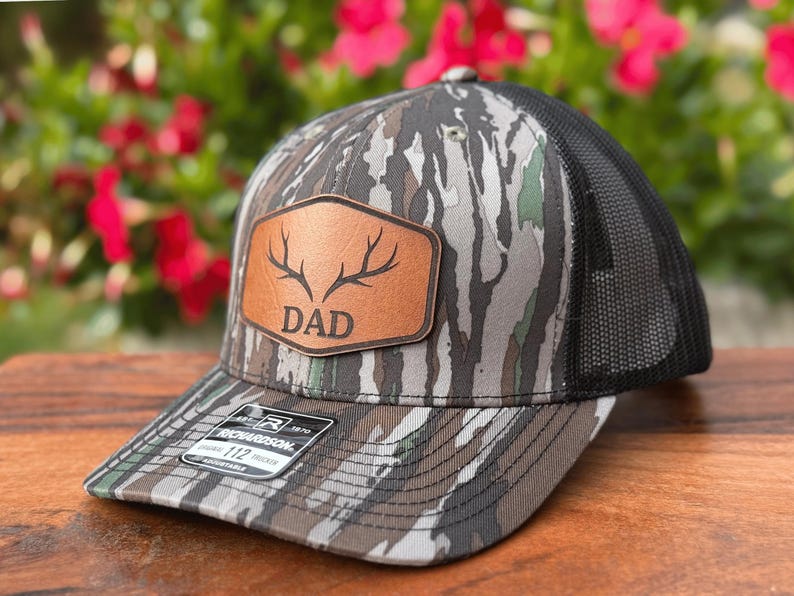 Pu&ograve; includere: Un cappellino trucker con motivo mimetico nei toni del marrone, grigio e verde e una parte posteriore in rete nera. Una toppa color cuoio sul davanti mostra un'illustrazione di corna di cervo sopra la scritta DAD. Un adesivo &egrave; presente sulla visiera.
