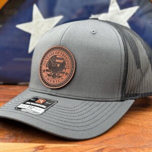May include: A grey baseball cap with a black mesh back and a circular brown leather patch on the front. The patch features an eagle graphic, stars, and the text 250 YEARS OF FREEDOM AMERICA. A black sticker on the brim displays the Richardson 112 logo.