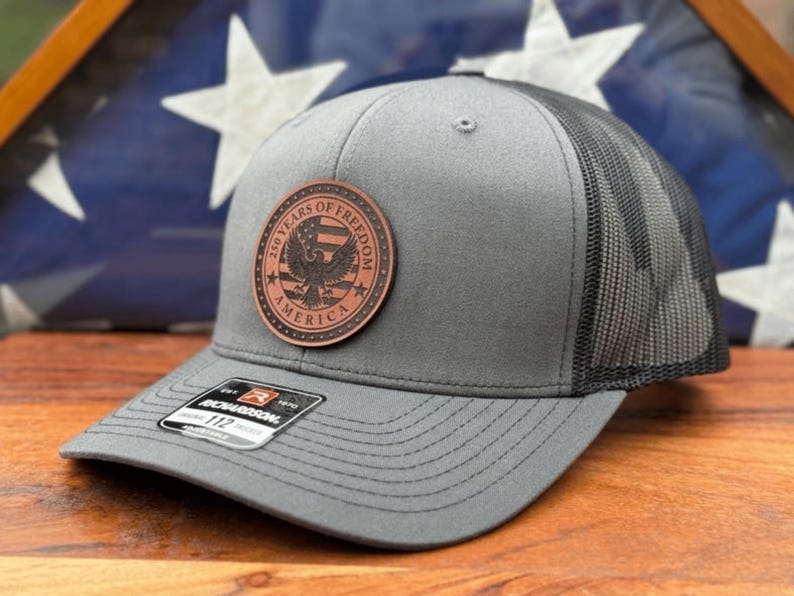 May include: A grey baseball cap with a black mesh back and a circular brown leather patch on the front. The patch features an eagle graphic, stars, and the text 250 YEARS OF FREEDOM AMERICA. A black sticker on the brim displays the Richardson 112 logo.