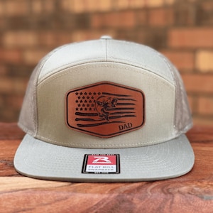 May include: A light gray snapback hat with a flat bill and a brown leather patch on the front. The patch features an engraved American flag design with a fish silhouette and the word DAD. A rectangular sticker is centered on the flat brim.