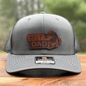 May include: Gray trucker hat with a brown leather patch that reads "BEST DAD" and features a fish graphic. The hat has a mesh back and a curved brim. The Richardson 112 adjustable hat is on a wooden surface.