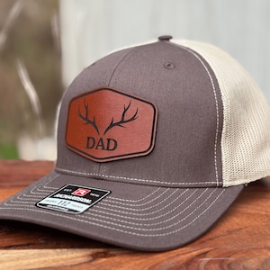 Pu&ograve; includere: Un cappellino trucker grigio antracite e beige con una toppa in pelle marrone sul davanti. La toppa a forma di esagono arrotondato presenta una grafica nera di corna di cervo sopra la parola DAD in un carattere grassetto senza grazie.