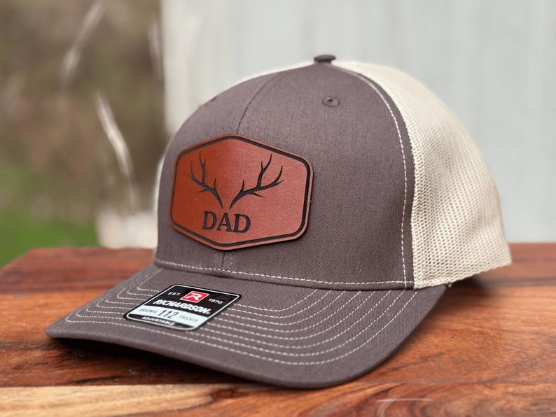 Pu&ograve; includere: Un cappellino trucker grigio antracite e beige con una toppa in pelle marrone sul davanti. La toppa a forma di esagono arrotondato presenta una grafica nera di corna di cervo sopra la parola DAD in un carattere grassetto senza grazie.