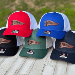 May include: Five different baseball caps with white mesh back panels and leather pennants with team names. The caps are red, blue, black, green, and navy blue. The pennants read "Cougars", "Wildcats", "Bulldogs", "Eagles", and "Panthers".