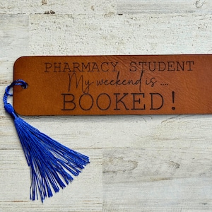 May include: Brown leather bookmark with a blue tassel. The bookmark has the text "Pharmacy Student My weekend is... BOOKED!" engraved on it.