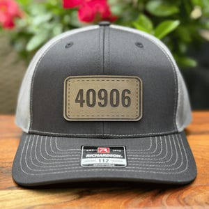 May include: A charcoal gray and white trucker hat with a leather patch displaying the number "40906". The hat has a curved brim and an adjustable strap. The brand tag "Richardson 112" is visible.
