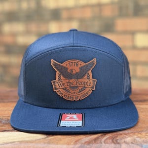 May include: A navy blue snapback hat featuring a brown leather patch on the front. The patch displays an eagle, the text 1776, We the People, and 250 YEARS OF LIBERTY. A rectangular sticker is centered on the flat bill with the text FLAT BILL SNAPBACK.