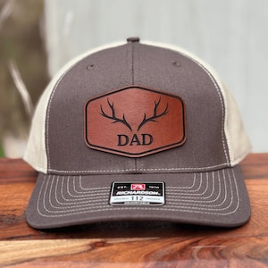 Pu&ograve; includere: Un cappellino trucker grigio antracite e beige con una toppa in similpelle marrone sul davanti. La toppa presenta un disegno inciso di corna di cervo sopra la scritta DAD. Un adesivo bianco e nero sulla visiera riporta la scritta RICHARDSON 112.
