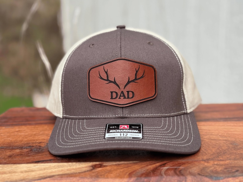 Pu&ograve; includere: Un cappellino trucker grigio antracite e beige con una toppa in similpelle marrone sul davanti. La toppa presenta un disegno inciso di corna di cervo sopra la scritta DAD. Un adesivo bianco e nero sulla visiera riporta la scritta RICHARDSON 112.