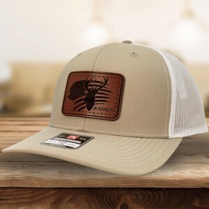 May include: A beige and white trucker hat featuring a rectangular brown leather patch on the front. The patch displays a silhouette of a deer head overlaid on an American flag design with the text JK RANCH printed in black lettering.