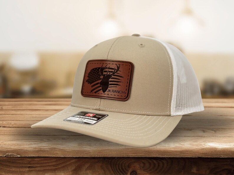 May include: A beige and white trucker hat featuring a rectangular brown leather patch on the front. The patch displays a silhouette of a deer head overlaid on an American flag design with the text JK RANCH printed in black lettering.