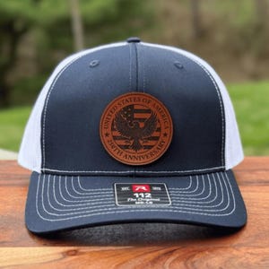 May include: A navy blue and white trucker hat featuring a circular brown leather patch on the front. The patch displays an eagle, a flag design, and the text United States of America 250th Anniversary. A sticker on the brim reads 112 The Original.