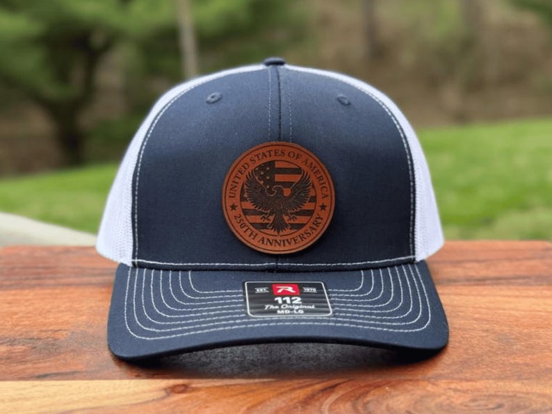 May include: A navy blue and white trucker hat featuring a circular brown leather patch on the front. The patch displays an eagle, a flag design, and the text United States of America 250th Anniversary. A sticker on the brim reads 112 The Original.