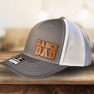 May include: A gray and white trucker hat with a brown leather patch that says "DANCE DAD".