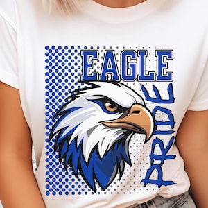 Könnte beinhalten: Weißes T-Shirt mit einem Grafikdesign eines Adlerkopfes in Blau und Weiß. Der Text "EAGLE PRIDE" ist blau. Das Design beinhaltet ein blaues Tupfenmuster, das einen auffälligen visuellen Effekt erzeugt.