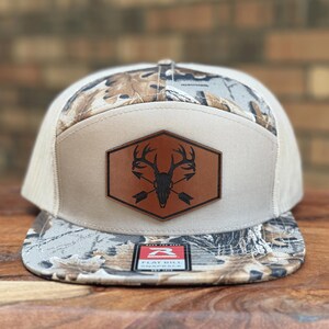 May include: A snapback hat featuring a camouflage pattern on the flat brim and upper panels, with a solid beige front panel. A brown hexagonal leather patch is centered on the front, displaying a black graphic of a deer skull with crossed arrows.