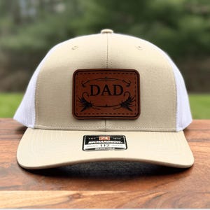 May include: Tan and white trucker hat with a brown leather patch that reads "DAD" in a stylized font. The patch also features fishing fly illustrations. The hat has a curved brim and a mesh back.