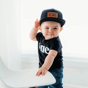 May include: A baby boy wearing a black baseball cap with a brown leather patch that says "BLAKE" on it. He is wearing a black t-shirt with the word "one" printed on it in white. He is also wearing blue jeans.