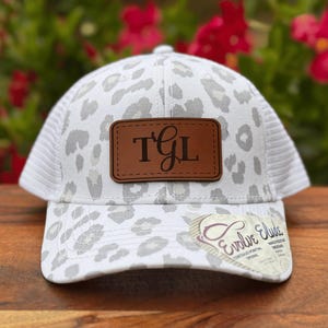 May include: White trucker hat with a gray leopard print pattern. The hat features a brown leather patch with the initials "TGL" and a curved brim. The back of the hat has a white mesh design. A tag with the text "Evolve Elusive" is attached.