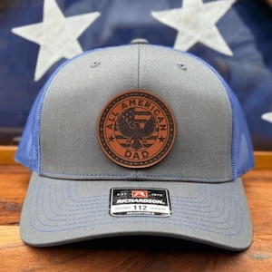 May include: A gray and blue baseball cap with a leather patch that reads "ALL AMERICAN DAD" with an eagle emblem. The cap has a mesh back and a curved brim. The brand tag "RICHARDSON 112" is visible.