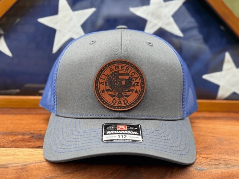 May include: A grey and blue trucker hat featuring a circular brown leather patch on the front. The patch displays an eagle, a flag, and the text ALL AMERICAN DAD. A black sticker on the brim reads RICHARDSON 112 ORIGINAL ADJUSTABLE.