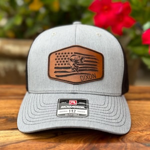 May include: A heather gray and black trucker hat featuring a brown leather patch on the front. The patch displays a laser-engraved American flag design with a fish silhouette and the text DIXON. A black sticker on the brim reads RICHARDSON 112.