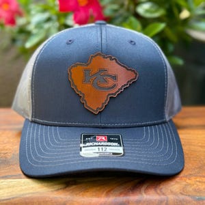 May include: Navy blue and gray baseball cap with a brown leather patch featuring a stylized "KC" logo. The cap has a curved brim and a mesh back. The Richardson 112 tag is visible on the front.