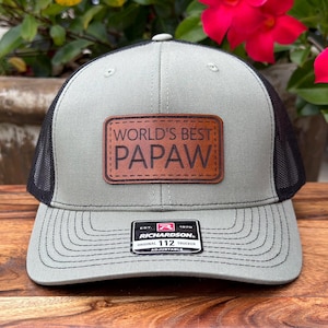 May include: A sage green and black trucker hat featuring a rectangular brown leather patch centered on the front panel. The patch is embossed with the text WORLD'S BEST PAPAW. A black and white Richardson 112 brand sticker is on the brim.