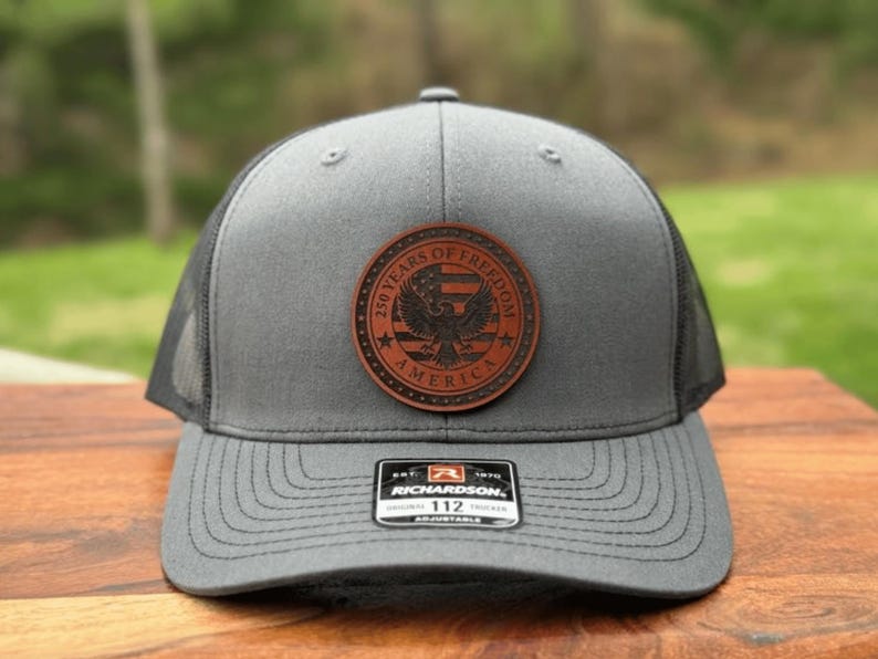 May include: A grey trucker hat with a black mesh back and a circular brown leather patch on the front. The patch features an eagle, an American flag, and the text 250 YEARS OF FREEDOM AMERICA. A black and white Richardson 112 sticker is on the brim.