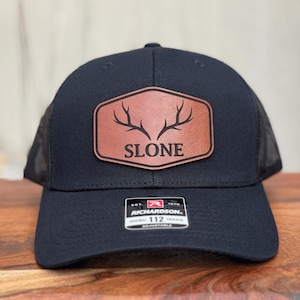 Pu&ograve; includere: Un cappellino trucker nero con una toppa in similpelle marrone sul davanti. La toppa &egrave; di forma esagonale con angoli arrotondati e mostra una grafica nera di corna di cervo sopra la scritta SLONE. Un adesivo Richardson 112 &egrave; sulla visiera.