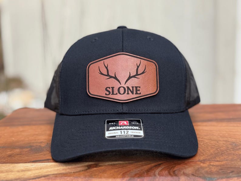 Pu&ograve; includere: Un cappellino trucker nero con una toppa in similpelle marrone sul davanti. La toppa &egrave; di forma esagonale con angoli arrotondati e mostra una grafica nera di corna di cervo sopra la scritta SLONE. Un adesivo Richardson 112 &egrave; sulla visiera.