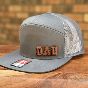 May include: A grey snapback trucker hat with a flat brim and a mesh back panel. The front panel features a brown leather-like patch with the word DAD embossed in a bold, block font. A small rectangular sticker is attached to the flat brim.