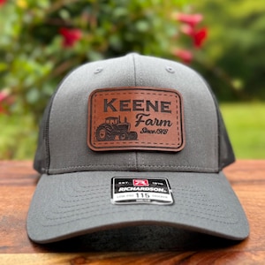 May include: A gray trucker hat with a black mesh back and a brown leather patch. The patch features the words "KEENE Farm Since 1918" and a tractor graphic. The hat has a curved brim and a Richardson 115 tag.