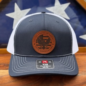 May include: A navy blue and white trucker hat featuring a circular brown leather patch on the front. The patch displays an eagle, a flag, and the text United States of America 250th Anniversary. A sticker on the brim reads R 112 The Original MD-LG.