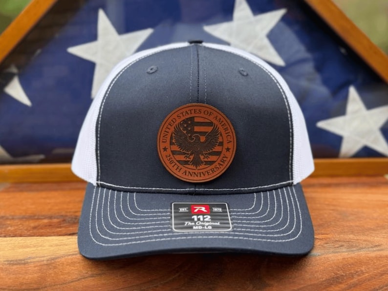 May include: A navy blue and white trucker hat featuring a circular brown leather patch on the front. The patch displays an eagle, a flag, and the text United States of America 250th Anniversary. A sticker on the brim reads R 112 The Original MD-LG.