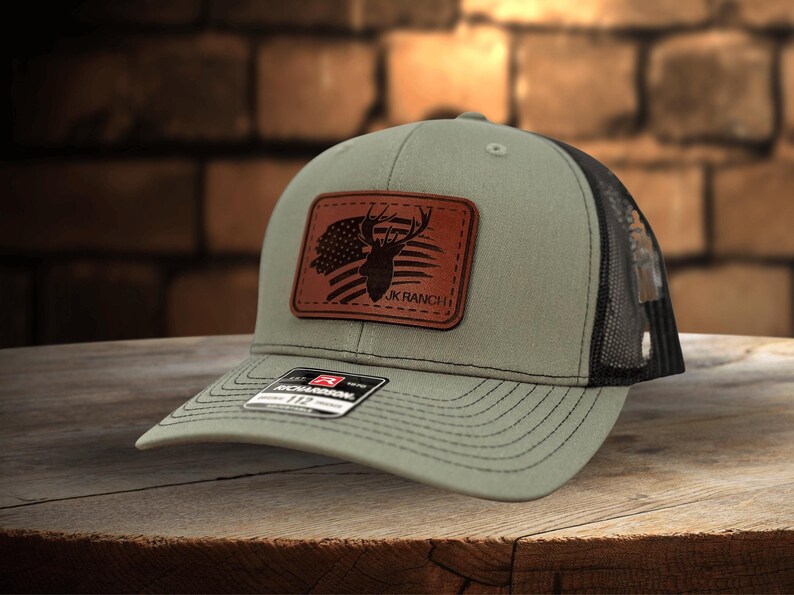 May include: An olive green baseball cap with a black mesh back and a rectangular brown leather patch on the front. The patch features a black laser-engraved graphic of a deer head silhouette overlaid on an American flag with the text JK RANCH.